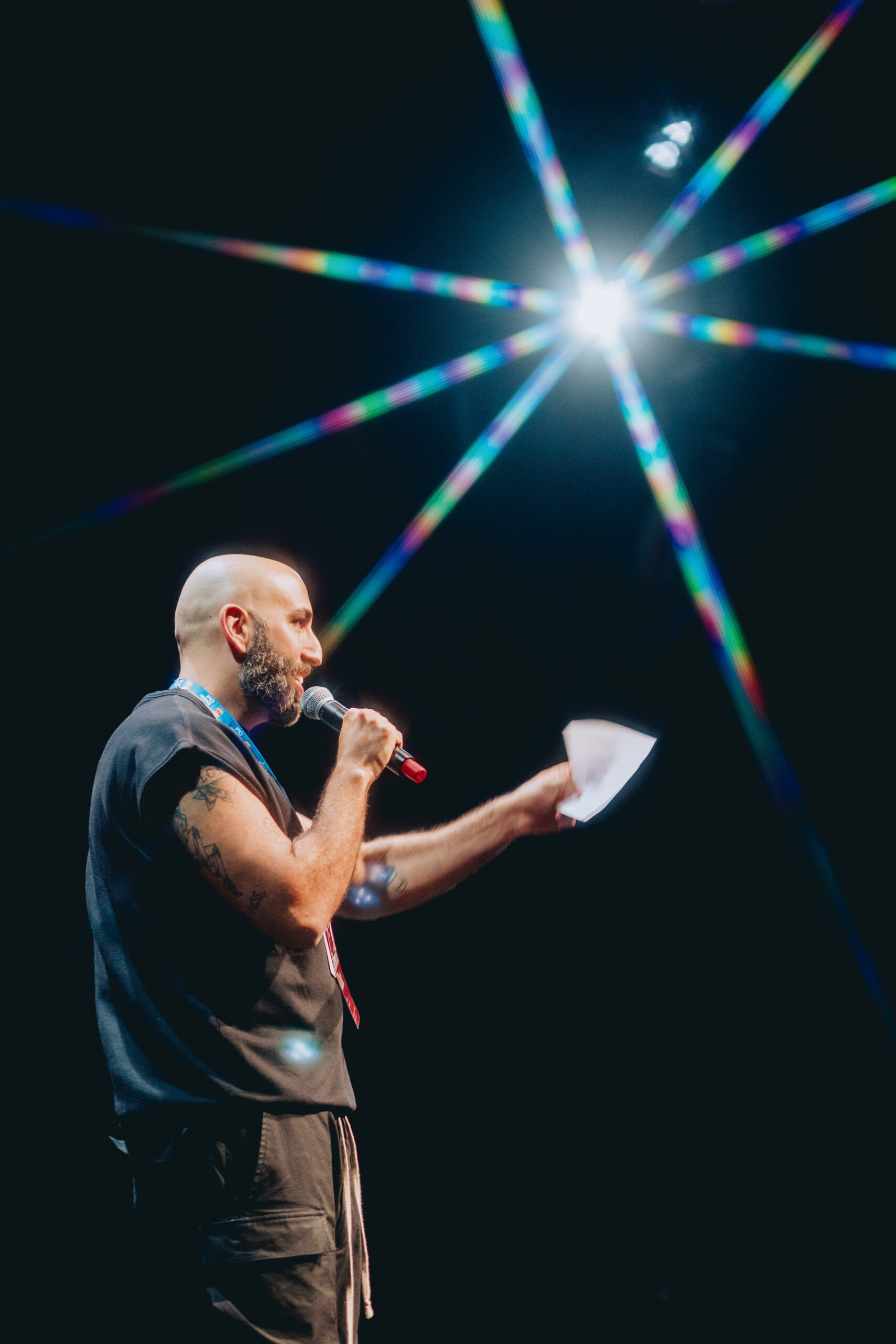 A person with a beard and tattoos speaks into a microphone on stage, holding papers. Bright stage lights create a starburst effect above them against a dark background.