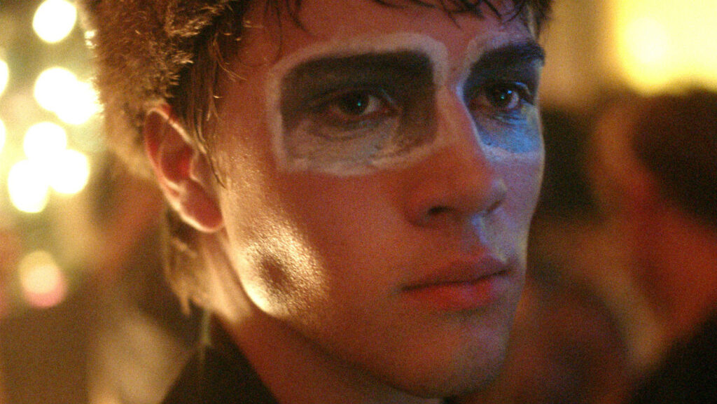 A young person with short brown hair stands in soft lighting, wearing dramatic black and white face paint around their eyes, looking off into the distance with a thoughtful expression.